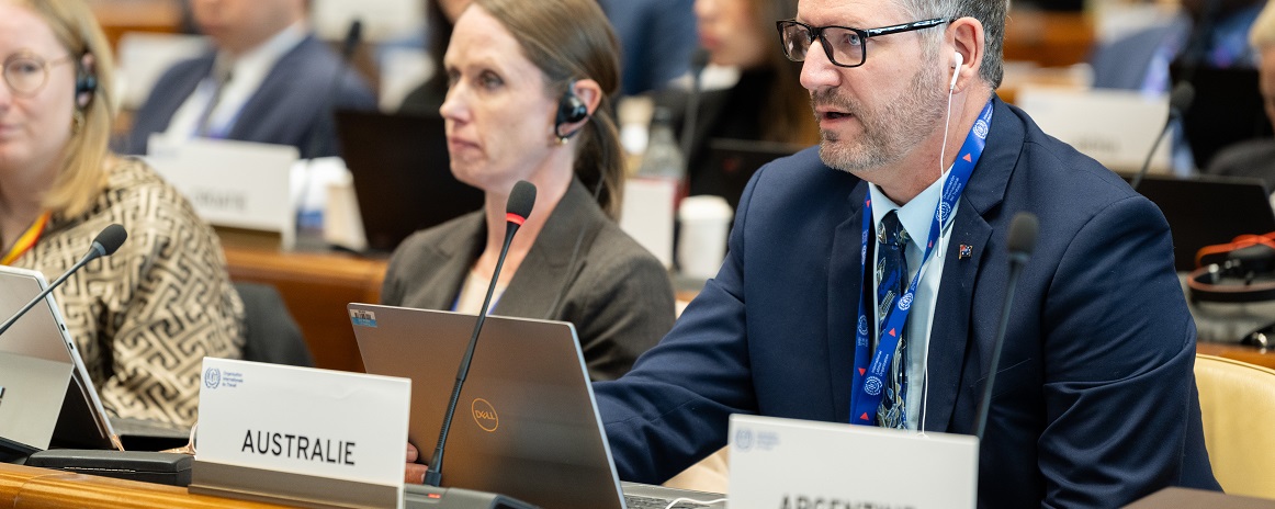 Delegate speaking at an international conference, seated behind a sign labeled “AUSTRALIE.” Wearing a suit and headset, with a laptop open. Other attendees are seated nearby, listening and using laptops. Signs for other countries, including “ARGENTINE,” are visible in the background.