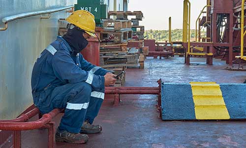 seafarer seated on piping on deck of large ship in work gear looking at his phone