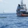 Ship with a boom pulled in close in order to recover the rust-coloured oil captured on the surface of the water