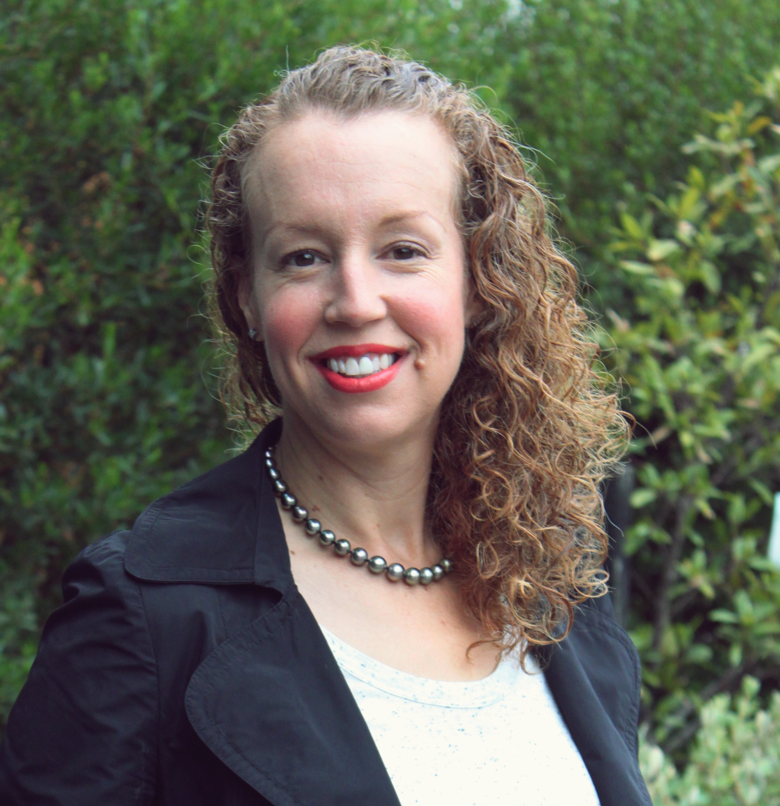 Donna standing outdoors smiling at camera. Donna has curly hair and wears a necklace with a navy blue jacket