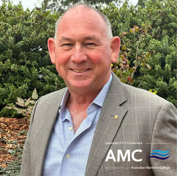 Mal is standing outside, smiling at the camera and wears a grey suit jacket and blue shirt