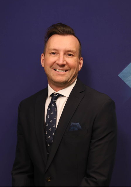 Greg Witherall smiles at the camera wearing a black suit, white collard shirt and navy blue tie