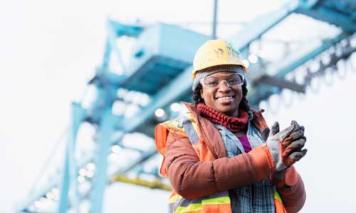 woman in hard hat, safety goggles, safety gloves and high-vis vest smiling with crane in background