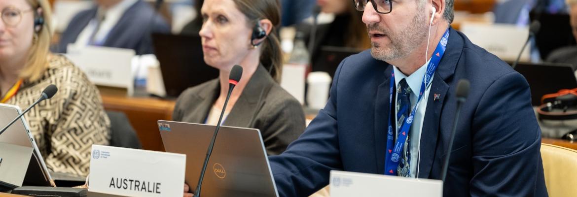 Delegate speaking at an international conference, seated behind a sign labeled “AUSTRALIE.” Wearing a suit and headset, with a laptop open. Other attendees are seated nearby, listening and using laptops. Signs for other countries, including “ARGENTINE,” are visible in the background.