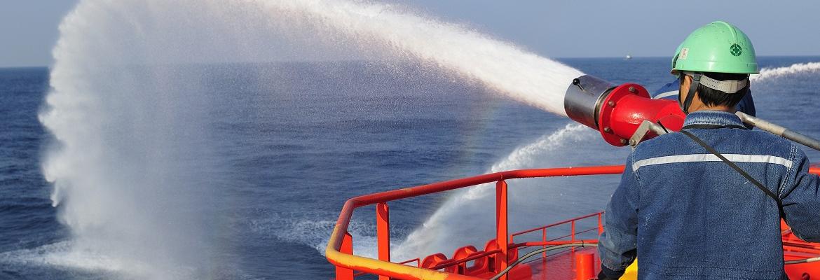 Person in a green helmet and blue coveralls operates a red fire monitor on a vessel, spraying a wide arc of water over the ocean. The equipment is bright red with orange railings, and a faint rainbow is visible in the mist.