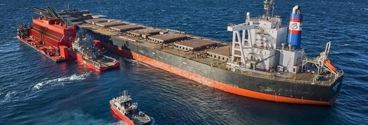 A large bulk carrier with an orange and black hull is underway at sea, accompanied by three red vessels nearby. A heavy industrial structure is secured on the carrier’s port side deck. The ship’s funnel features a bold red, white, and blue stripe design.