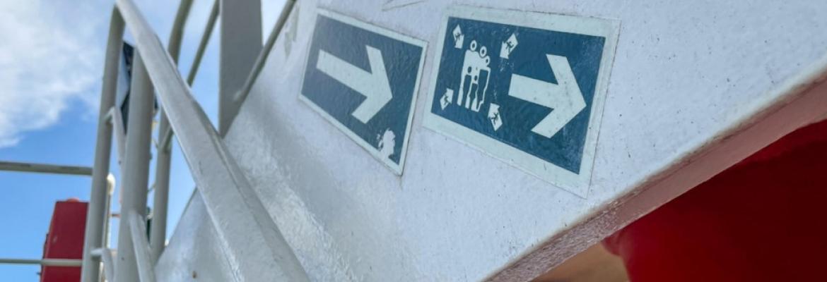 Two blue directional signs on a ship’s stairway point to the right, indicating the route to the assembly station. Blue sky and railing visible in the background.