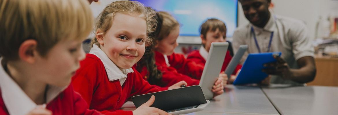 A group of students aged between five and seven sitting around a table and being shown how to use tablets by two teachers (male and female). 