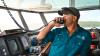 Person holding a marine radio to their mouth. They are wearing a blue 'crew' shirt with a hat and sunglasses on their head. They are on a boat’s bridge, surrounded by navigation equipment and a fire extinguisher in the background.