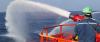 Person in a green helmet and blue coveralls operates a red fire monitor on a vessel, spraying a wide arc of water over the ocean. The equipment is bright red with orange railings, and a faint rainbow is visible in the mist.