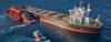 A large bulk carrier with an orange and black hull is underway at sea, accompanied by three red vessels nearby. A heavy industrial structure is secured on the carrier’s port side deck. The ship’s funnel features a bold red, white, and blue stripe design.